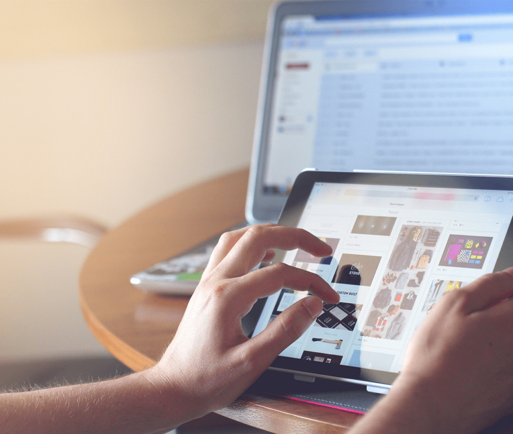 Person scrolls on a tablet as their laptop sits in the background and open to their emails