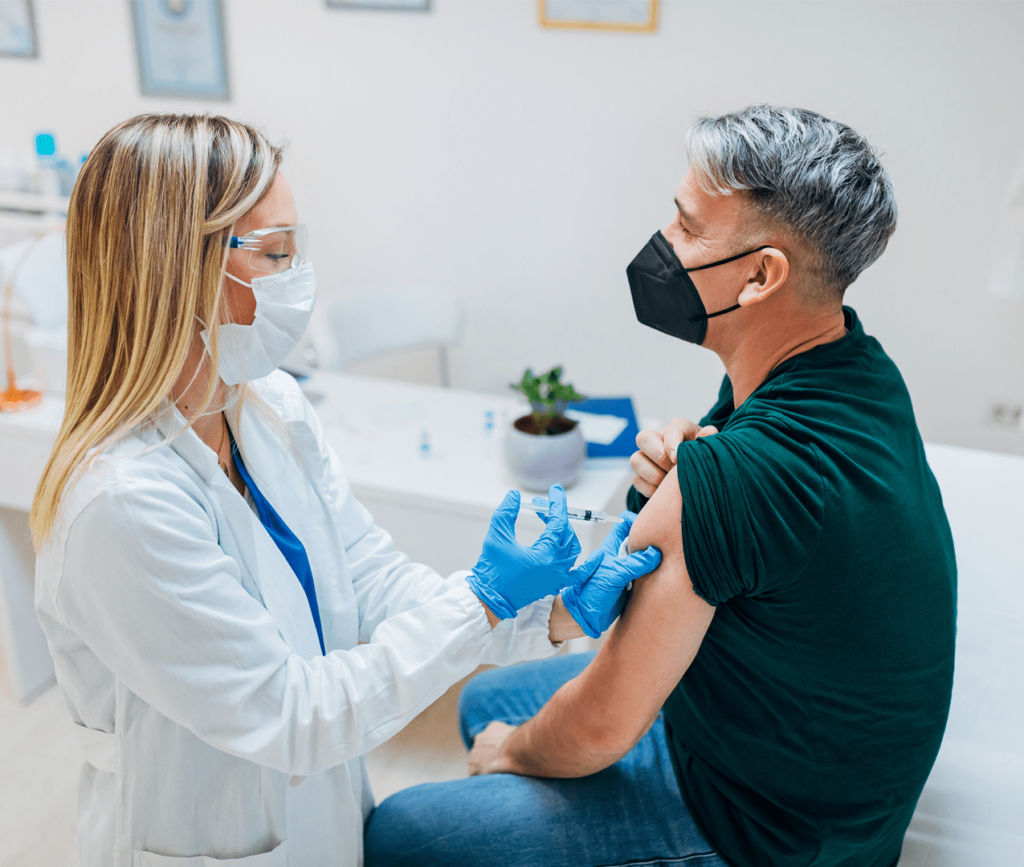 Male, middle age patient sitting on a doctors exam table being administered a shot by a female doctor, each with COVID masks on.