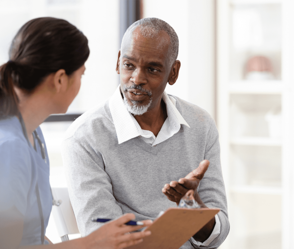 Senior man sitting with a doctor discusses his health as the doctor listens and takes notes on a clipboard