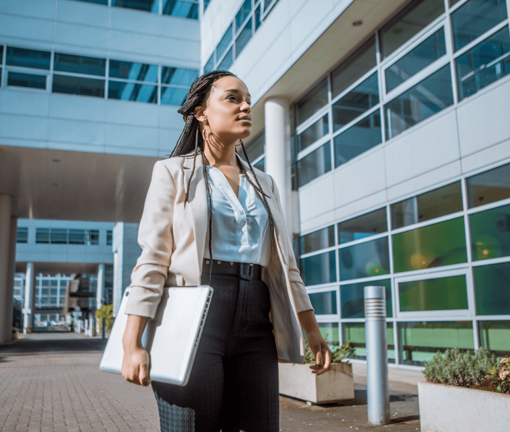 Young, female business professional standing outside of an office building, holding her laptop as she looks up at the office.