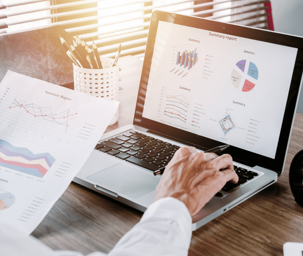 Business executive looking at a printed report of different metrics and graphs as he holds the paper with one hand and types on his laptop with the other. His laptop showing a dashboard of more metrics than in the printed report.