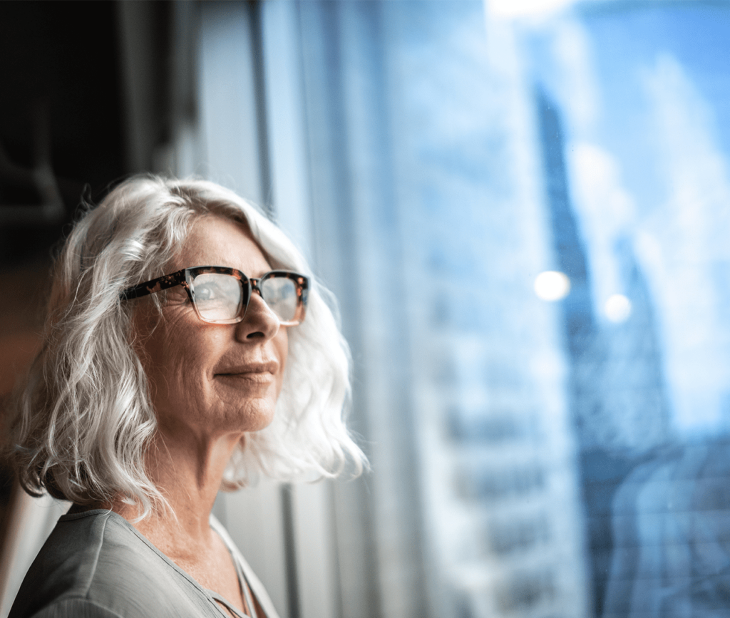 Elderly woman stares out of the window of a high-rise building on a sunny day.