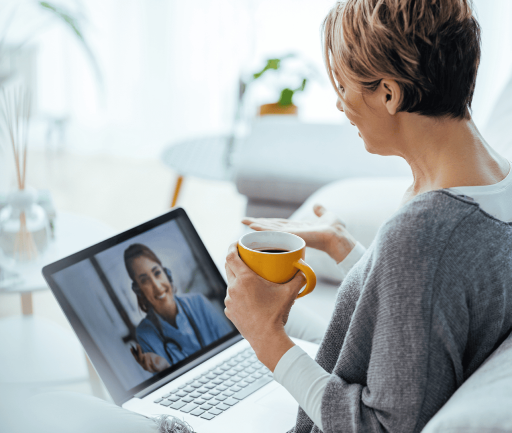 Older woman sits with her laptop and coffee and has a video call with her friendly doctor.