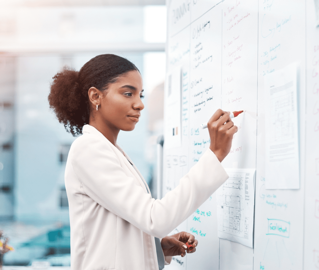 Young female data scientist writes notes and wireframe on a whiteboard as she diagrams out a process.