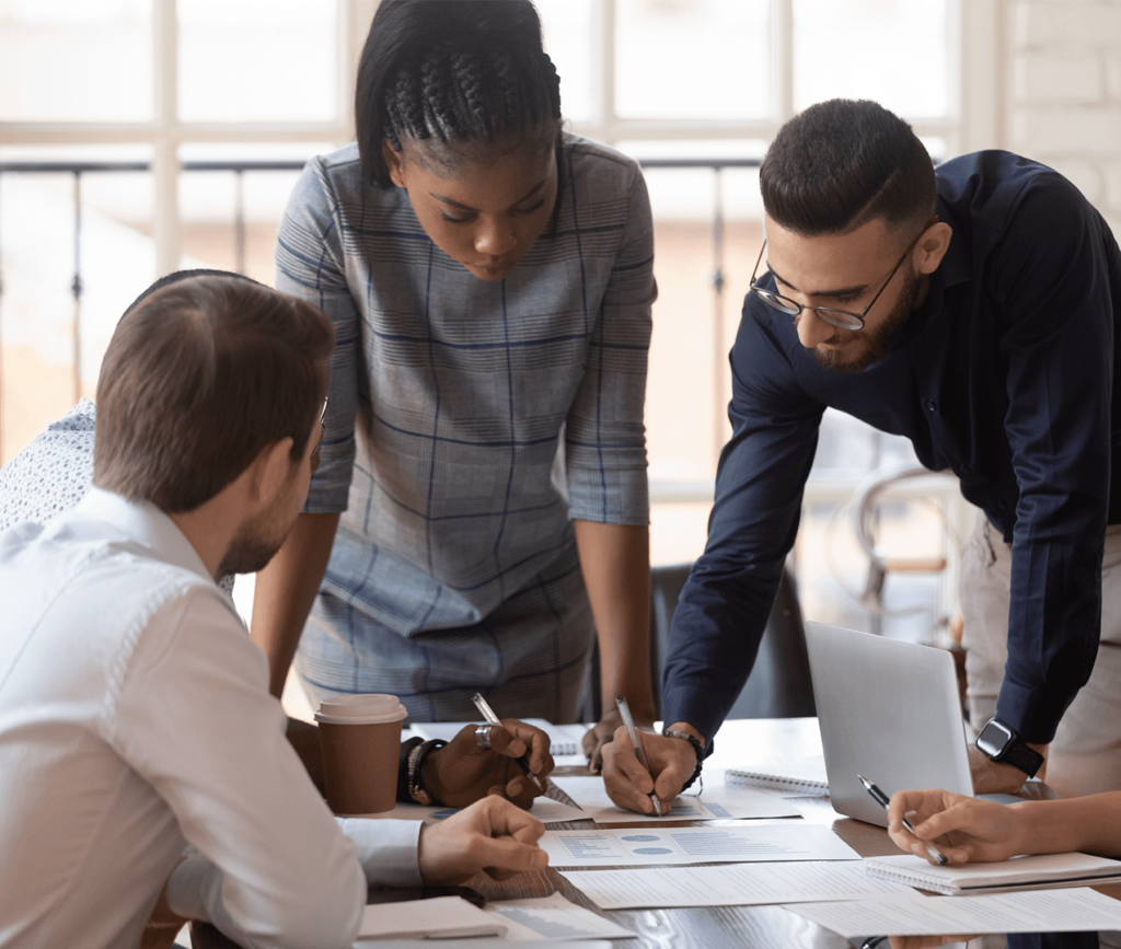 Business team of 5 diverse individuals reviewing and making notes on several reports scattered across the boardroom table.