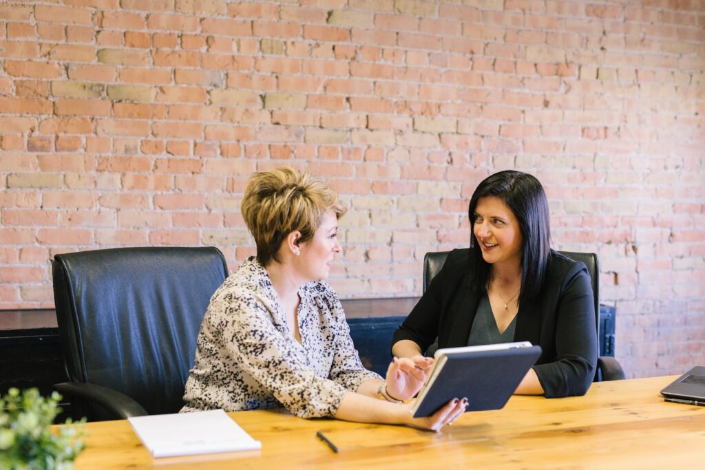 Two businesswomen speak at a conference room table as one woman holds a tablet for discussion.