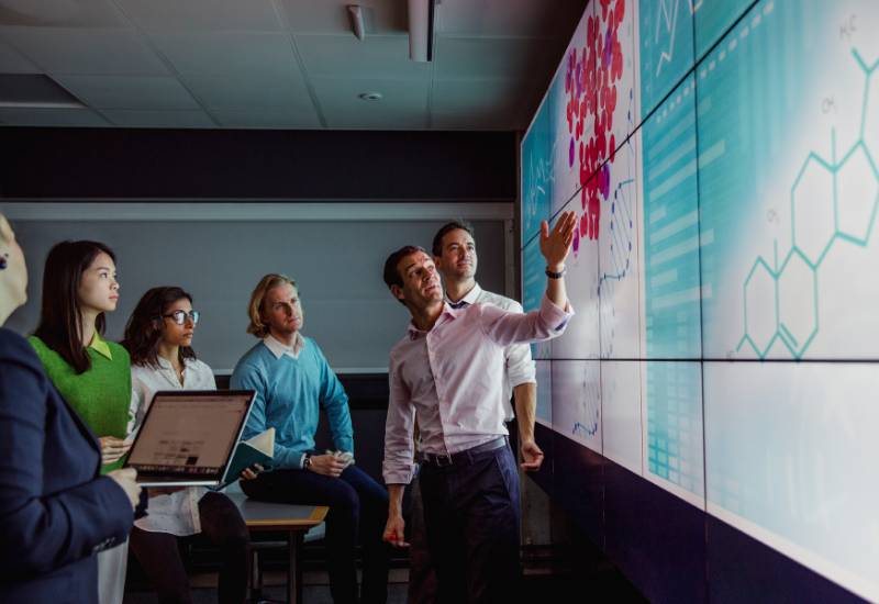 Male science researcher leading a group meeting on drug development with a large screen behind him displaying chemical compounds, DNA helix and data charts.