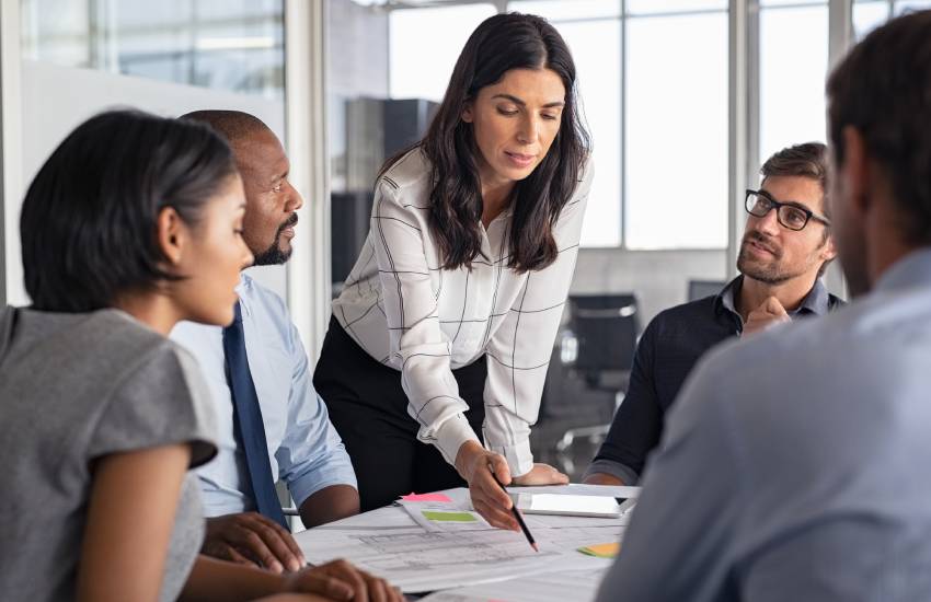 Business meeting being lead by a standing middle-aged woman pointing to reports scattered on the table with her pencil.