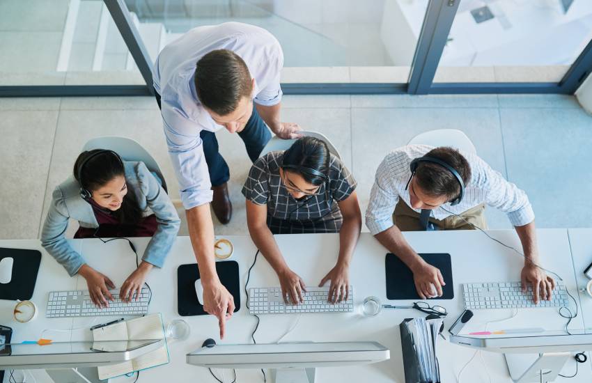 Top view of customer service help desk workers with headsets on and manager assisting by pointing to something of importance on the computer.