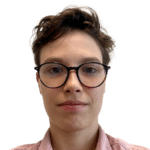 A close-up of a person with short brown hair and round dark-rimmed glasses, wearing a light pink collared shirt.